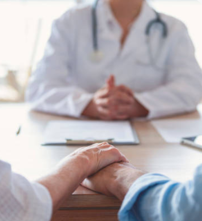 Doctor with stethoscope sits behind a desk, two people hold hands across the table during a consultation.